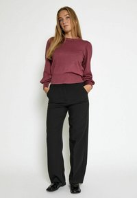 Mujer de pie contra un fondo blanco, llevando una camiseta de manga larga color burdeos, pantalones anchos negros y mocasines negros, con las manos en los bolsillos.