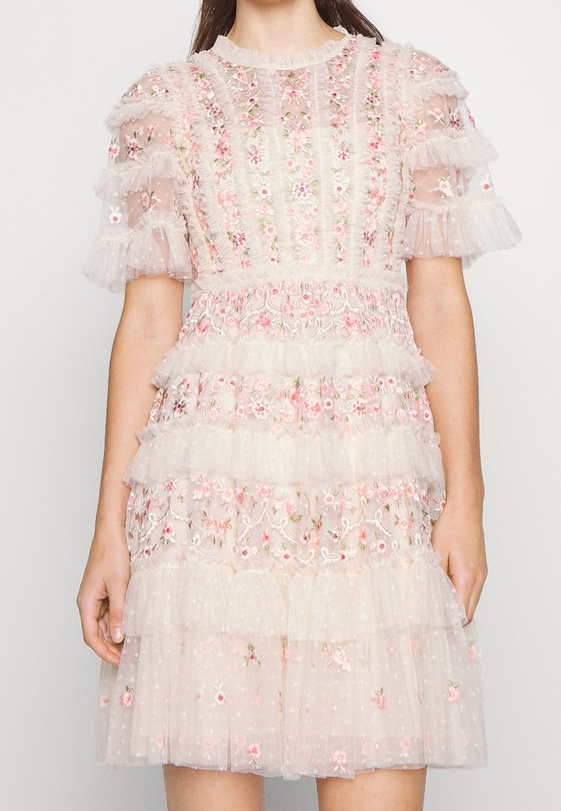 Femme portant une robe blanche transparente avec une broderie florale rose et des volants à étages aux manches courtes et à la jupe.