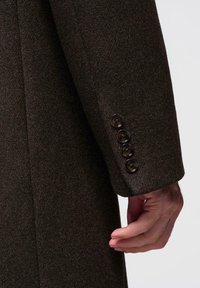 Hnědý vlněný kabát s hladkou texturou, se čtyřmi černými knoflíky na manžetě a upraveným rukávem, který vychází z bundy.
