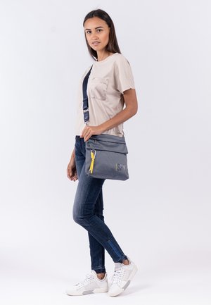 Gray messenger bag with textured fabric, yellow zipper pull, and logo detail. Worn crossbody, paired with a beige top and blue jeans.