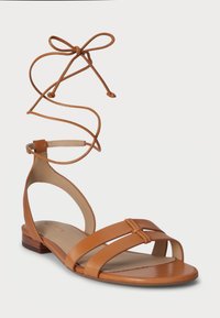 Καφέ δερμάτινο σανδάλι με λεπτές, σταυρωτές λωρίδες και δέσιμο στον αστράγαλο. Διαθέτει επίπεδη ξύλινη σόλα και λείο, στρογγυλό σχέδιο στη μύτη.