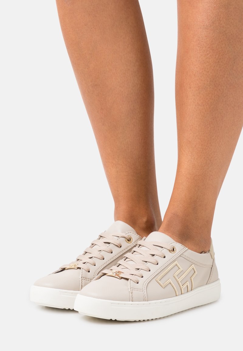 Baskets en cuir beige à texture lisse, avec des accents en métal doré et une semelle en caoutchouc blanche. Détail du logo visible sur le côté.