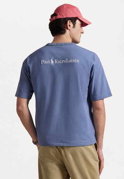 T-shirt bleu délavé à manches courtes en tissu texturé, avec un logo brodé blanc "Polo Ralph Lauren" au dos. Coupe décontractée.