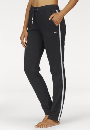 Woman wearing black sweatpants with white side stripes and drawstring waist, standing barefoot with one hand in pocket.