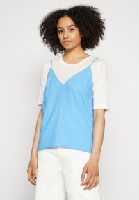Camisola de linho azul com alças finas, usada sobre uma blusa de algodão de manga curta branca. O conjunto apresenta um corte descontraído e texturas suaves.