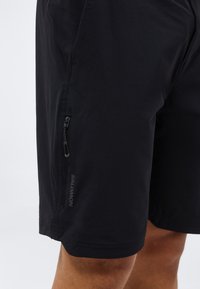 Μαύρα αθλητικά σορτς με πλαϊνή τσέπη με φερμουάρ και λογότυπο Salomon, φορεμένα από ένα άτομο με ορατό πόδι κάτω από τα σορτς.