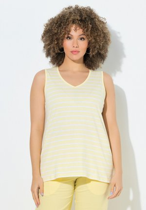 Vrouw met krullend haar die grote oorbellen draagt, een mouwloos geel-wit gestreept topje en gele broek, staand tegen een witte achtergrond.