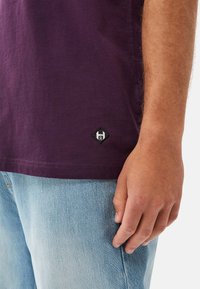 Lilla bomulds t-shirt med en lille sort og hvid logo-patch ved kanten. Skjorten er parret med lyseblå denim jeans.