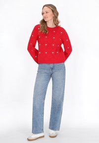 Pulover roșu tricotat cu modele de inimi și texturi de diamant. Asortat cu blugi amplați de culoare albastru deschis și adidași albi.