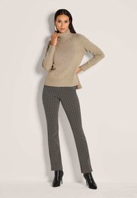 Frau in beigem Rollkragenpullover, schwarz-weißen gemusterten Hosen und schwarzen, hochhackigen Stiefeln, die vor einem schlichten, hellen Hintergrund posiert.