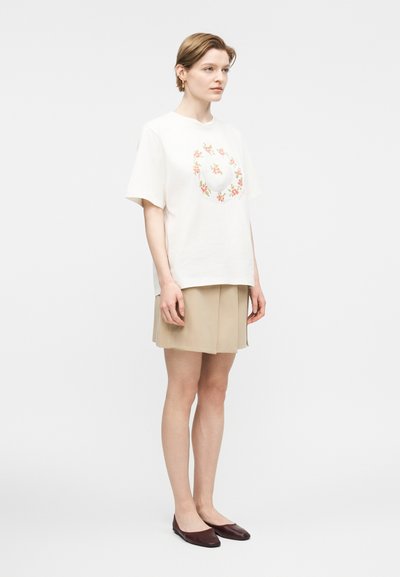 Γυναίκα στέκεται φορώντας λευκό κοντομάνικο T-shirt με σχέδιο στεφάνι με λουλούδια, μπεζ πλισέ φούστα και σκούρα καφέ μπαλαρίνες, μπροστά σε απλό φόντο.