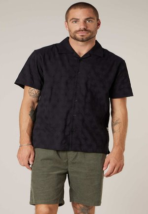 Man met kort haar en baard, gekleed in een zwart overhemd met korte mouwen en olijfgroene korte broek, staand tegen een effen achtergrond.