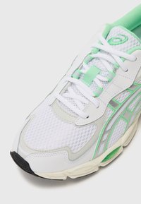White mesh sneaker with green accents, featuring a gray overlay, padded collar, and textured sole. Includes white laces and branding detail.