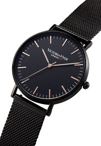 Hodinky s černým síťovaným páskem, kulatým černým ciferníkem a růžově zlatými indexy a ručičkami. Na ciferníku je nápis "VICTORIA-HYDE LONDON" a minimalistický design.