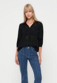 Black knit top with a v-neck and long sleeves, featuring cut-out designs and textured patterns, paired with blue striped denim pants.