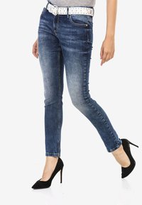 Jeans skinny bleu avec un design délavé, ornés d'une ceinture cloutée blanche, associés à des talons hauts en daim noir. Texture en denim lisse, coupe slim.