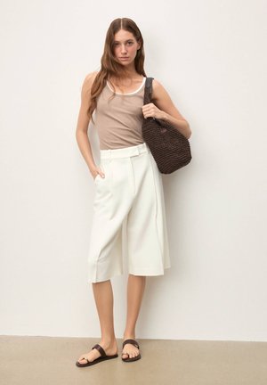 Pantaloni albi cu croială largă, un top bej fără mâneci cu margini albe și sandale maro. Modelul poartă o geantă maro texturată, împletită.