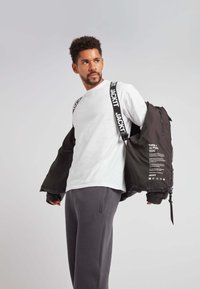 Giacca nera con bretelle a logo bianco, indossata aperta su una camicia bianca e pantaloni grigi. Presenta polsini elasticizzati e un'etichetta stampata sul retro.
