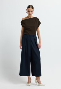 Top marrone senza spalline con design drappeggiato, abbinato a pantaloni a gamba larga blu. La modella indossa tacchi alti beige e orecchini rotondi.
