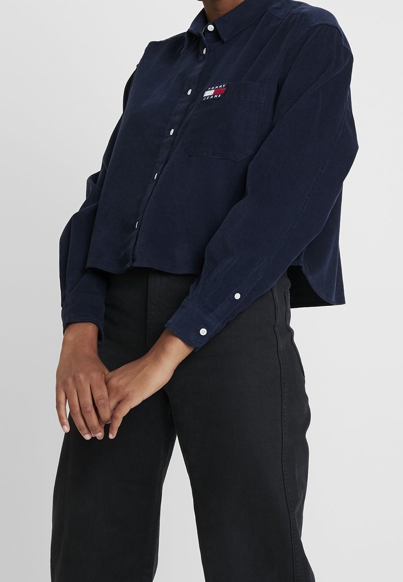 Camicia di velluto a coste blu navy con colletto a punta, taschino frontale, bottoni bianchi e logo ricamato sul petto. Design crop.
