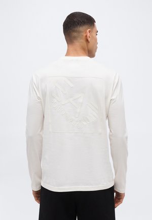 Mees, kes kannab valget pikkade varrukatega särk, mille tagaküljel on reljeefne Armani logo, seisab seljaga vaataja poole, taust on helehall ja ühtlane.