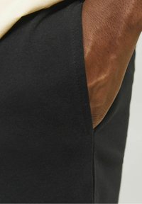 Tissu noir avec une texture côtelée, gros plan sur la zone de la poche, laissant entrevoir une teinte de peau. Design simple et ajusté. Pas de motifs ni de détails matériels.