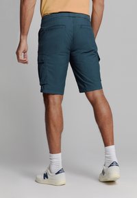 Teal cargoshorts met een elastische tailleband, zijzakken en achterzakken. G gedragen met witte sokken en New Balance sneakers.