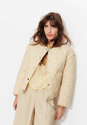 Femme aux longs cheveux bruns porte une veste matelassée crème sur un haut brodé jaune clair et un pantalon beige, debout devant un fond blanc.