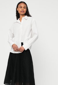 Cămașă albă cu guler, având mâneci lungi și o croială lejeră, asortată cu o fustă neagră plisată care ajunge până la mijlocul gambei.