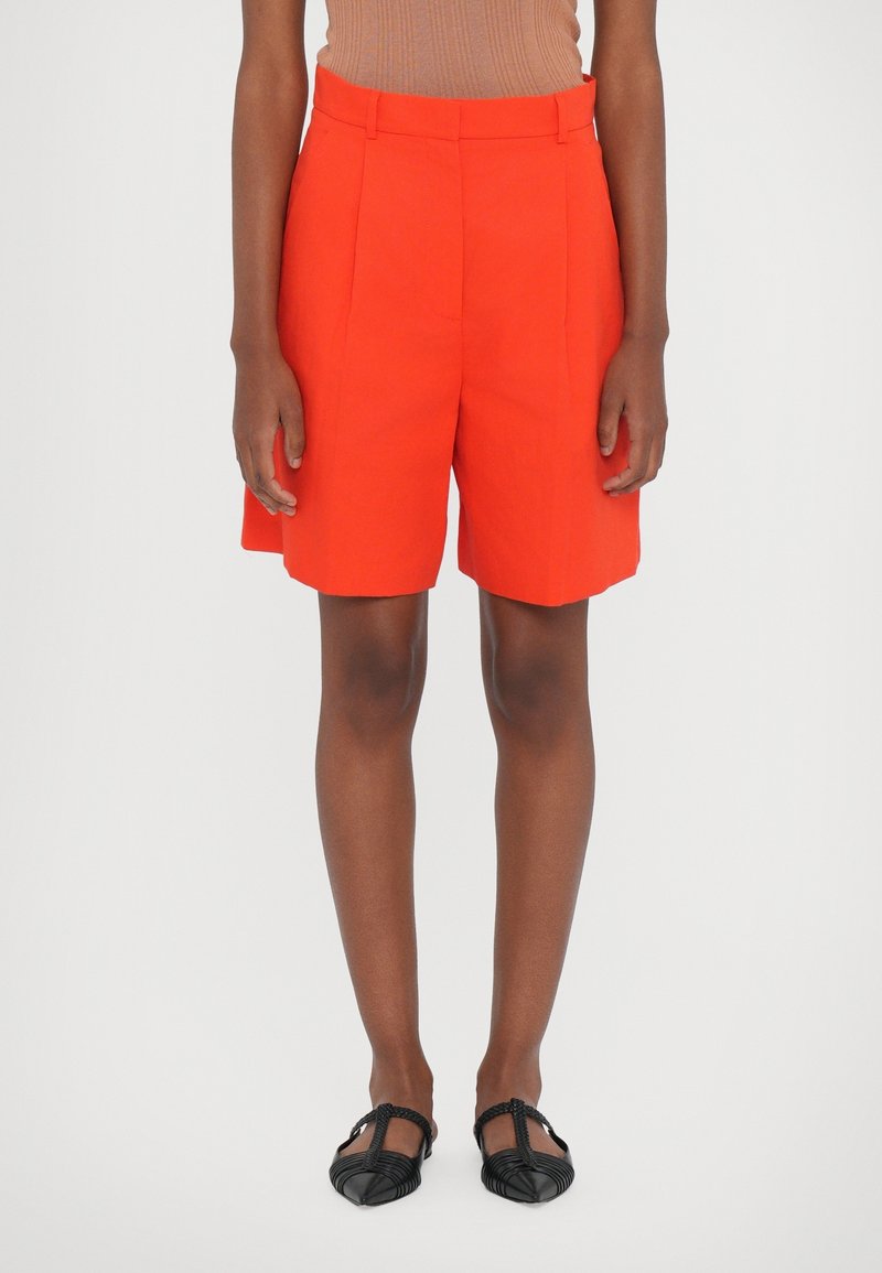 Shorts rouges sur mesure à taille haute avec des plis, fabriqués dans un tissu léger. Portés avec des chaussures plates noires au design tissé.