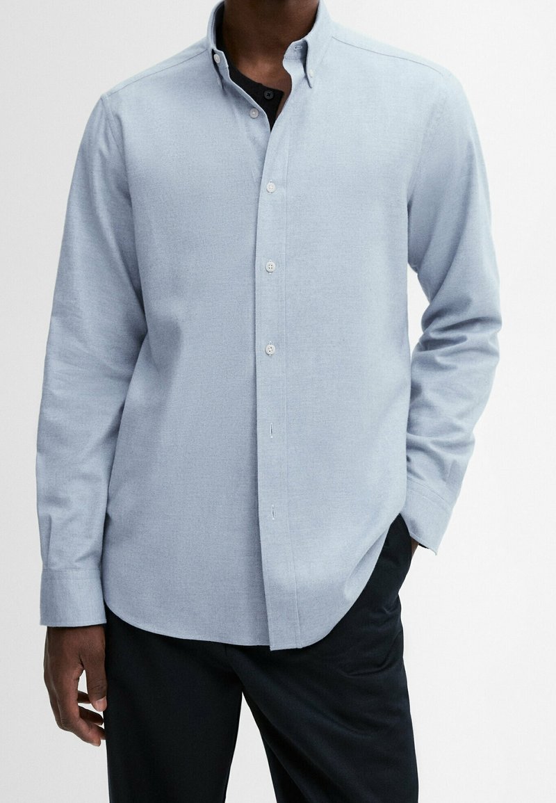 Homme portant une chemise boutonnée à manches longues bleu clair par-dessus un t-shirt noir avec un pantalon noir, une main dans la poche, sur un fond uni.