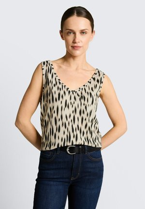 Femme aux cheveux foncés portant un haut beige sans manches à taches noires et un jean bleu foncé, debout les mains dans le dos.