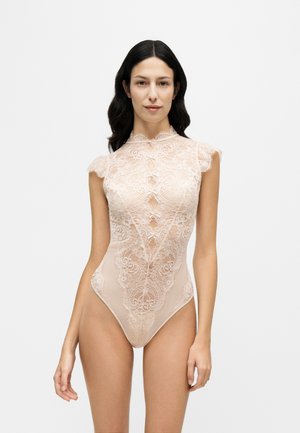 Una modelo posa sobre un fondo blanco liso, con un body de encaje rosa pálido, de cuello alto y manga corta.
