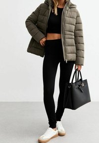 Puchowa oliwkowa kurtka z kapturem, czarny crop top, czarne legginsy, białe sneakersy z beżowymi podeszwami i usztywniona czarna torebka.