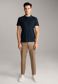 Marineblaues Poloshirt mit strukturiertem Muster, kombiniert mit hellbeigen Chinos und weißen Sneakers. Das Modell steht vor einem neutralen Hintergrund.
