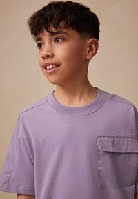 Fioletowa koszulka z krótkim rękawem wykonana z miękkiej bawełny, z naszywaną kieszonką na lewej piersi oraz okrągłym dekoltem. Gładka faktura.