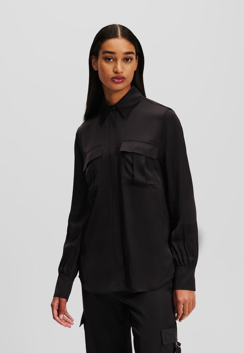 Schwarzes Satin-Hemd mit langen Ärmeln, Kragen, zwei Brusttaschen und gerafften Ärmeln für Textur. Schlankes, minimalistisches Design.