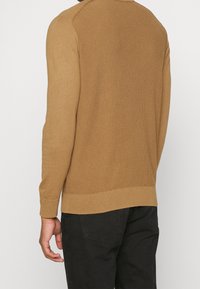 Ljusbrun stickad tröja med ribbat mönster, rund halsringning och åtsittande ärmar, kombinerad med svarta byxor, vilket visar en avslappnad stil.