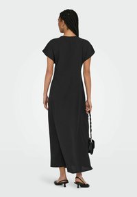 Robe maxi noire à manches courtes, silhouette fluide, tissu léger et détails au dos. Associée à un petit sac noir et des sandales.