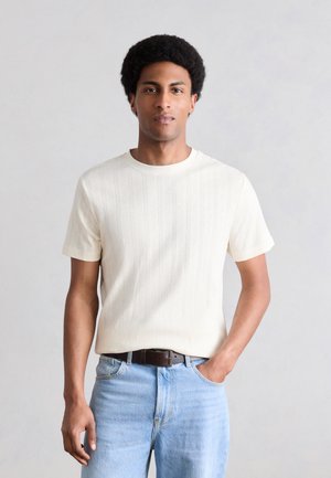 Muž s krátkymi kučeravými vlasmi, oblečený v bielej štruktúrovanej košeli s krátkym rukávom zastrčenej do svetlomodrých džínsov s hnedým opaskom, stojaci pred jednoduchým pozadím.