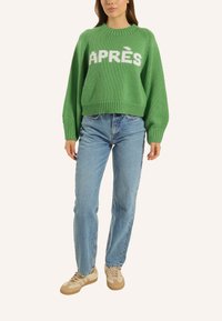 Grüner, gestrickter Pullover mit weißem "APRÈS"-Text, rundem Kragen und geripptem Saum. Kombiniert mit hellblauen Jeans und beigen Sneakern.