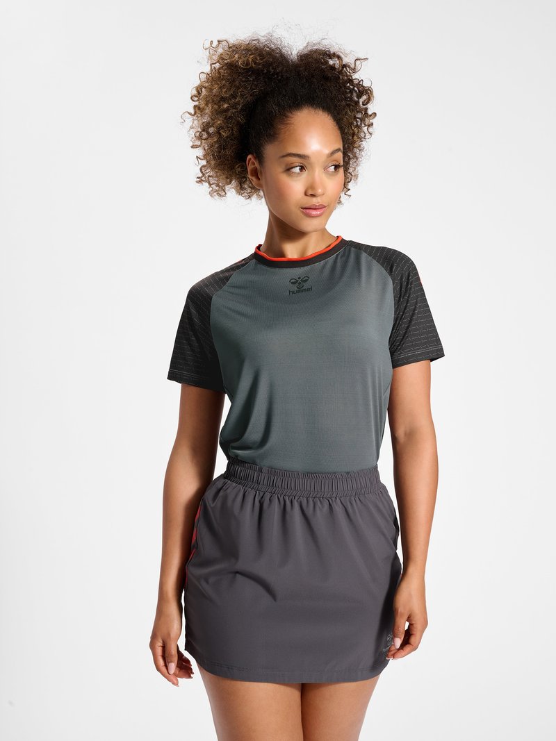 Vrouw met krullend haar die een grijs-zwart sportshirt en een donkergrijze rok met elastische taille draagt, staande tegen een witte achtergrond.