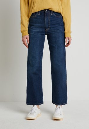 Jean en denim bleu foncé à taille haute et jambes larges, avec des détails de couture subtils. Porté avec un sweat-shirt jaune et des baskets blanches.
