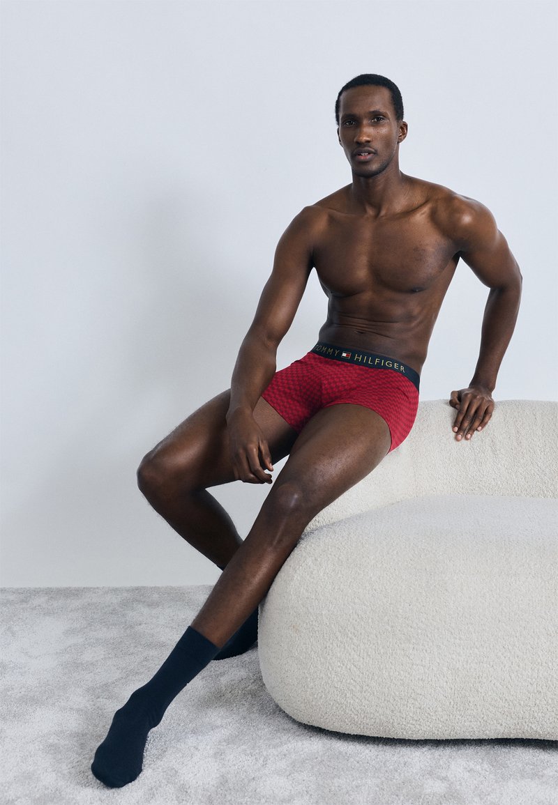 Rote Boxershorts mit Muster und einem schwarzen elastischen Bund mit Logo. Schwarze Socken. Model sitzt auf einer texturierten hellen Oberfläche.