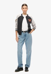 Veste bomber noire et grise avec poignets rayés, arborant un logo sur la poitrine. Associée à un jean bleu clair et des chaussures noires. Texture lisse.