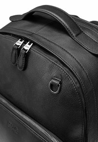 Czarny skórzany plecak o fakturowanym wykończeniu, z podwójnymi zamkami błyskawicznymi, górną rączką i okrągłym metalowym detalem z przodu.