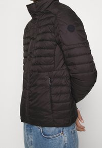 Černá péřová bunda s vysokým límcem, horizontálním prošíváním, kapsou na levé straně a kruhovým logem na rameni. Hladká textura.