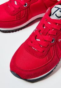 Scarpe da ginnastica rosse con un mix di tessuto liscio e pelle scamosciata testurizzata. Design con lacci, suola bianca e battistrada nera. Dettaglio di branding ben visibile.