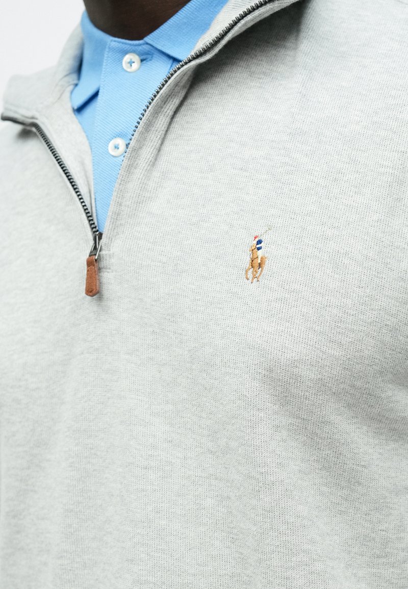 Hellgrauer Reißverschluss-Pullover aus strukturiertem Stoff, mit einem blauen Polo-Kragen darunter und einem kleinen gestickten Logo eines Pferdes.