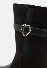 Bottines en daim noir avec une boucle en argent en forme de cœur et un panneau élastique pour plus d'élasticité. Accents en cuir noir lisse sur la bande.